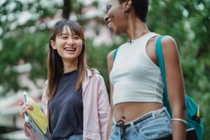 Two happy college girls laughing together on campus, one holding a laptop from her college tech kit