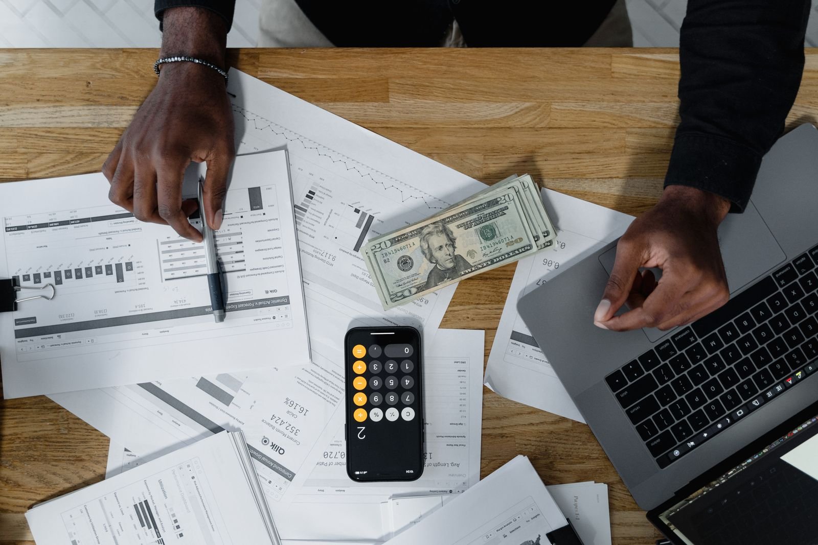 Budgeting by hand with a laptop, calculator, and cash on a wooden desk.