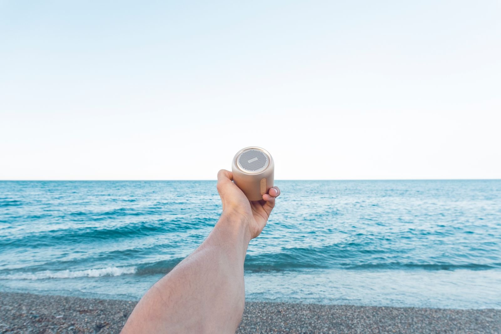 Hand holding a small portable speaker in front of the sea on a bright summer day.