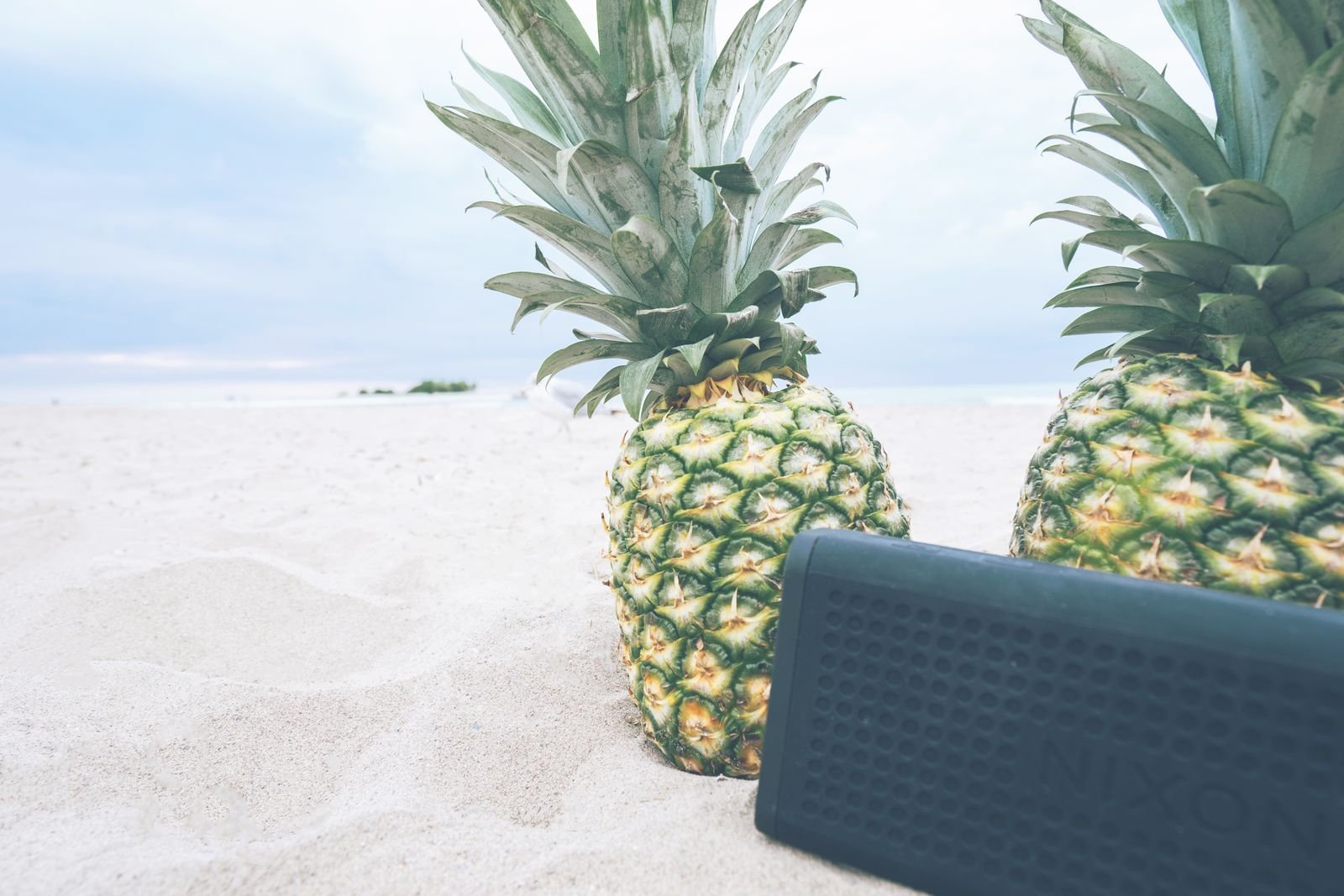 A rugged waterproof bluetooth speaker on a sunny sandy beach beside two pineapples.